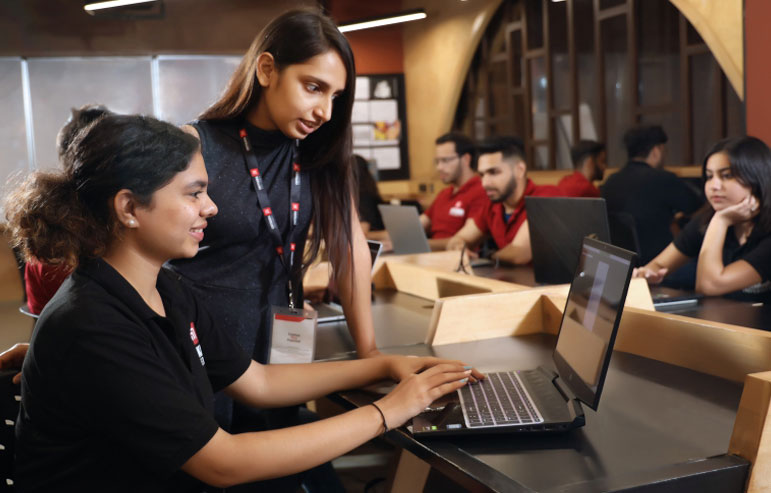 The image shows the students of chitkara university in black t-shirts and some in red. One works on the laptop, and the other stands by her side.
