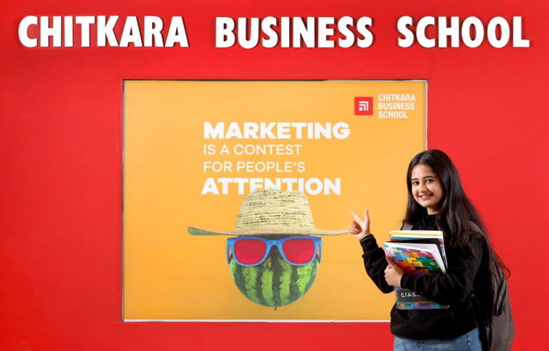 The illustration with a red background shows the text "CHITKARA BUSINESS SCHOOL" in bold white text in the centre. Below this text is an orange square box with a chitkara university logo and" MARKETING is A CONTEST FOR PEOPLE'S ATTENTION" in white. Below this text is a watermelon wearing a sun hat and sunglasses. On the left side of the image, there is a girl with open hair wearing a black sweatshirt, holding books and pointing towards an orange box.