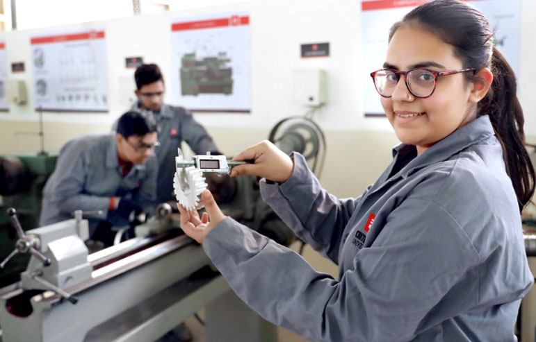 The image with blurred background shows the engineering students at chitkara univeristy. Two are working on the machines, and the other is holding some equipment and smiling.