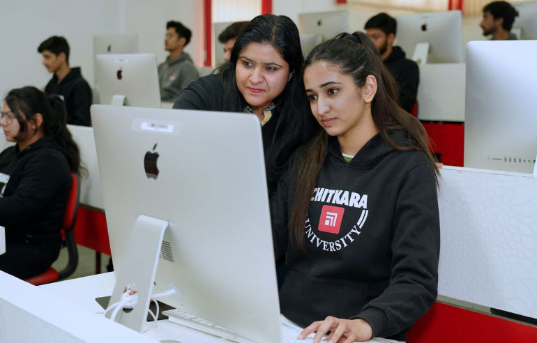 The image shows the students of chitkara university in black sweatshirts working on the desktops. There is also a teacher who teaches something to the student.