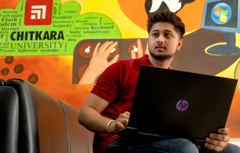 An image with colourfull background with a chitkara university logo shows a student in red t-shirt holding a laptop.