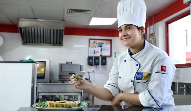 A culinary student in professional chef uniform preparing a gourmet dish in a training kitchen, representing hands-on learning and global careers in culinary management.